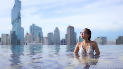 attractive asian female in infinity rooftop pool of luxury bangkok hotel enjoying sun, cityscape in background