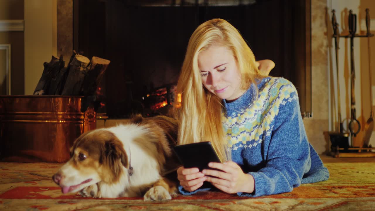 벽난로 에 누워있는 백인 젊은 여성, 태블릿을 즐기십시오. 그녀의 에는 개가 있습니다.
