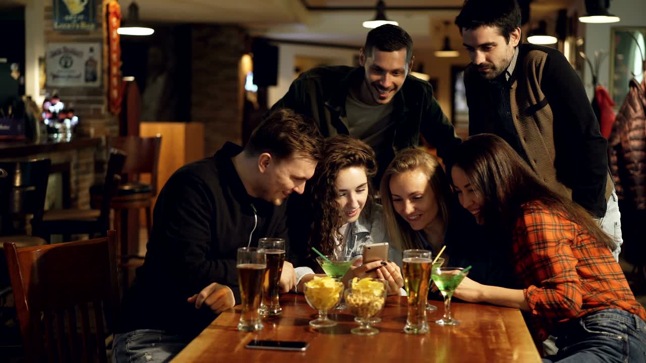 Happy students are watching photos in smartphone, discussing them and laughing while sitting at table in bar. Snacks and drinks in foreground, chairs and tables in background.