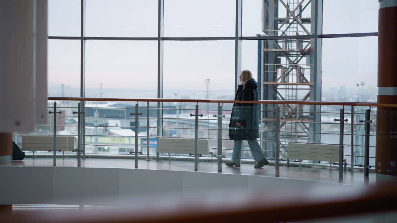 검은색 재을 입은 젊은 여성이 현대 쇼핑몰에서 철 철로에 손을 들고 고 있으며, 뒤에서 흐릿한 도시와 움직이는 자동차가 보이고 있으며, 큰 유리 창문은 도시 풍경을 보여줍니다.