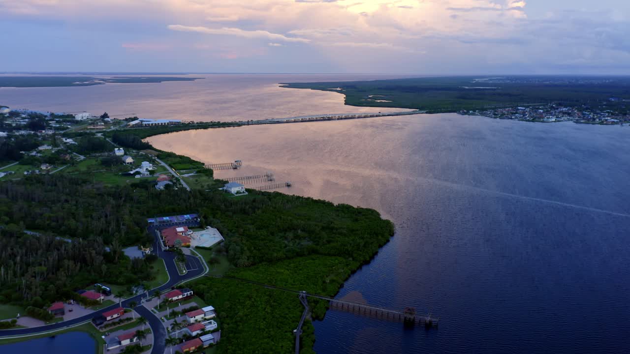 A peaceful coastal town sits along curving waterways that lead into a vast bay, where the sunset sky glows over still water, blending human architecture and tranquil natural beauty