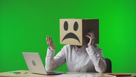 Person with a cardboard box head using a phone in a office setting