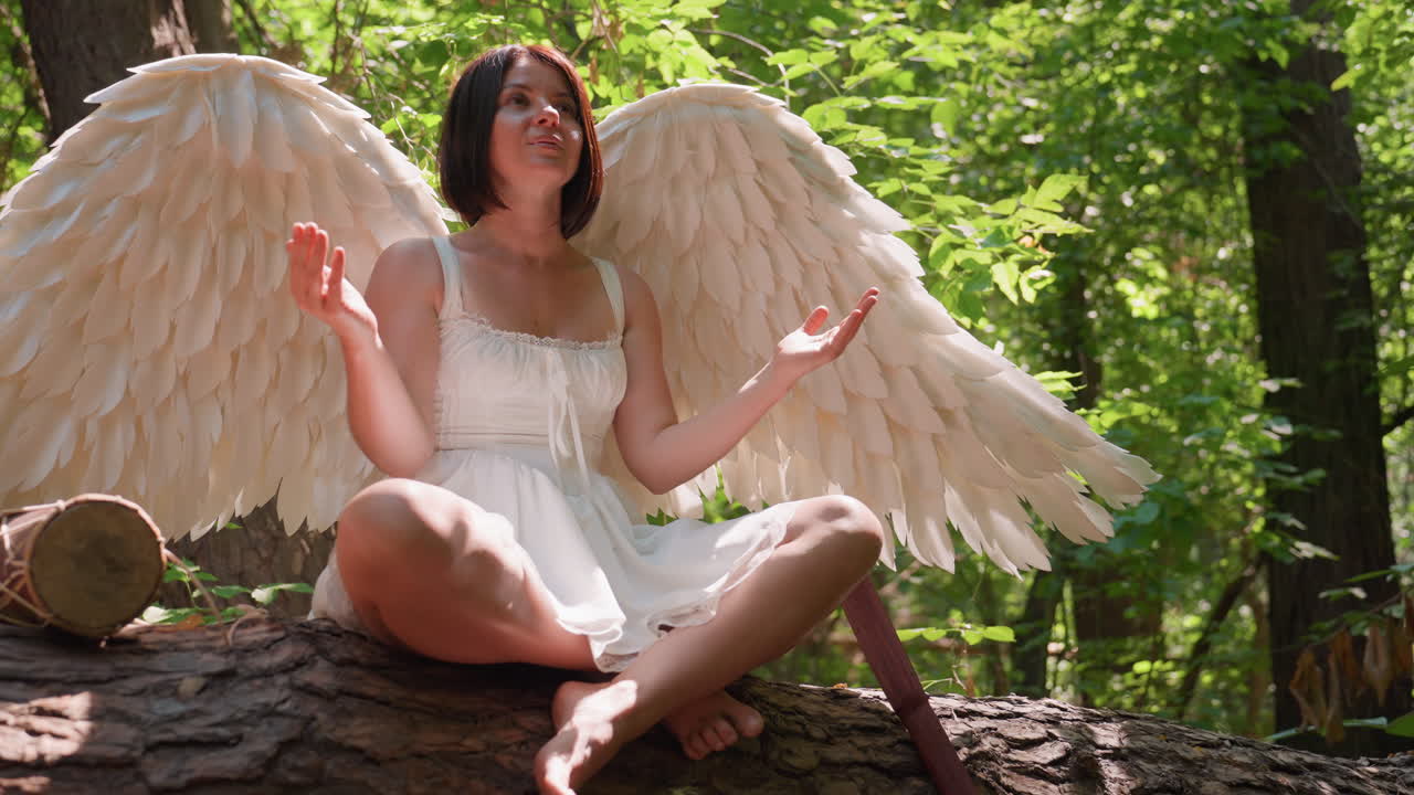 Young woman dressed in white angel costume sitting cross leg on fallen forest tree meditating peacefully under sunlight surrounded by green leaves, with talking drum and wooden staff by side