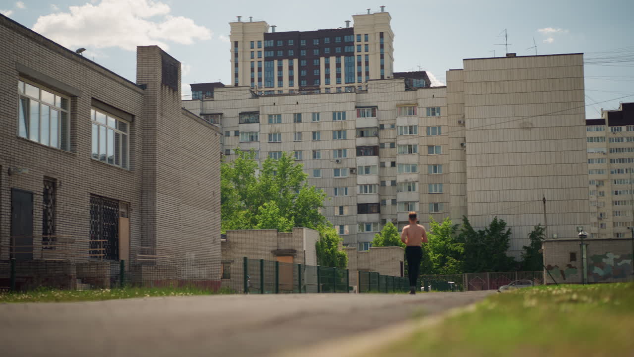 건물 사이의 안뜰 길을 조깅하는 셔츠를 입지 않은 백인 남성, 꾸준한 쿨다운 보폭, 콘크리트 캠퍼스와 녹색 지대, 따뜻한 햇빛과 편안한 호흡, 사려 깊은 훈련 일시 중지