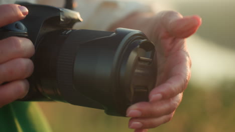 close up of photographer gently placing lens cap onto camera lens in soft warm outdoor light, hands visible with natural fingernails, green outfit slightly visible, serene grassy field in background