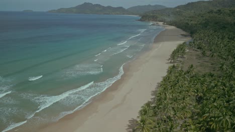 Waves-wash-over-sunny-beach-by-forest-in-sunny-Philippines,-aerial-pan