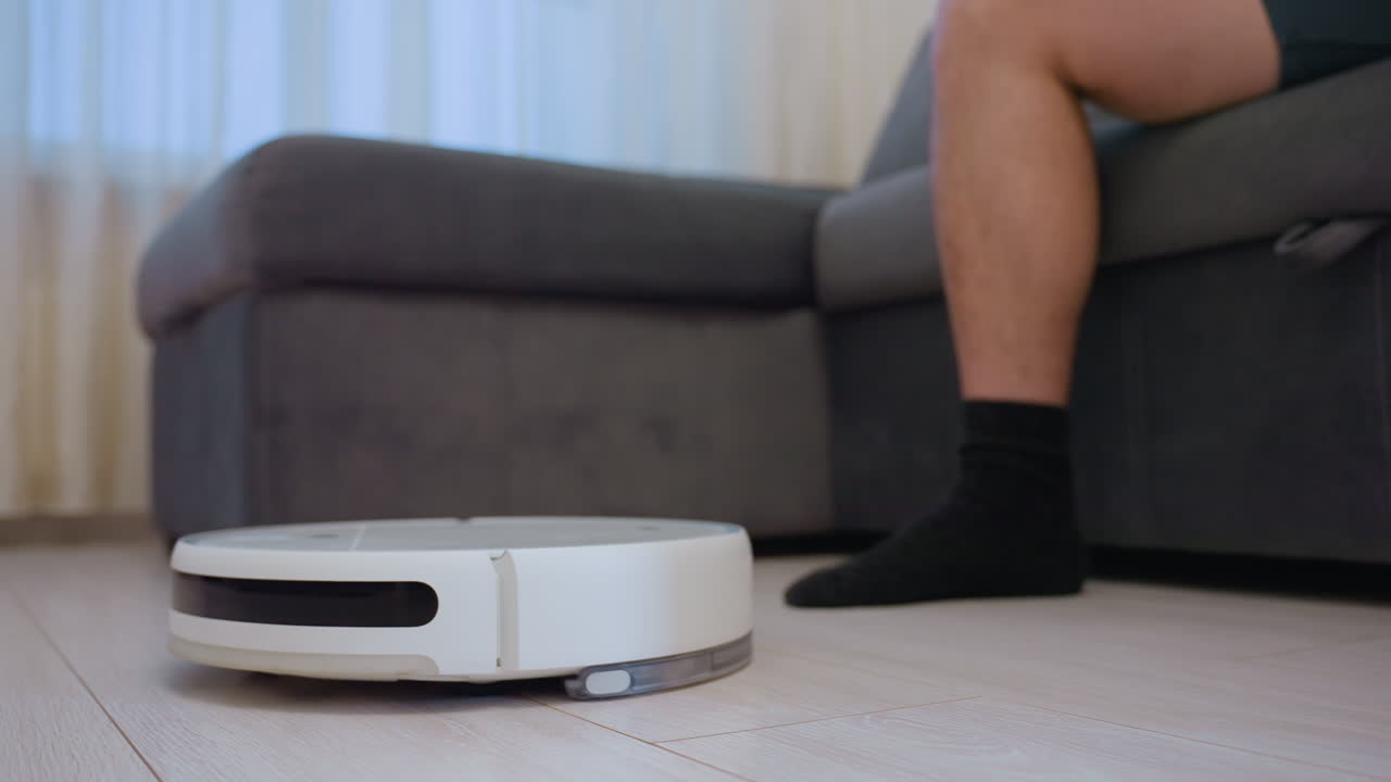 Vacuum robot transports headset across floor toward man seated on sofa using phone, as he collects headset and vacuum cleaner turns away, illustrating smart home convenience
