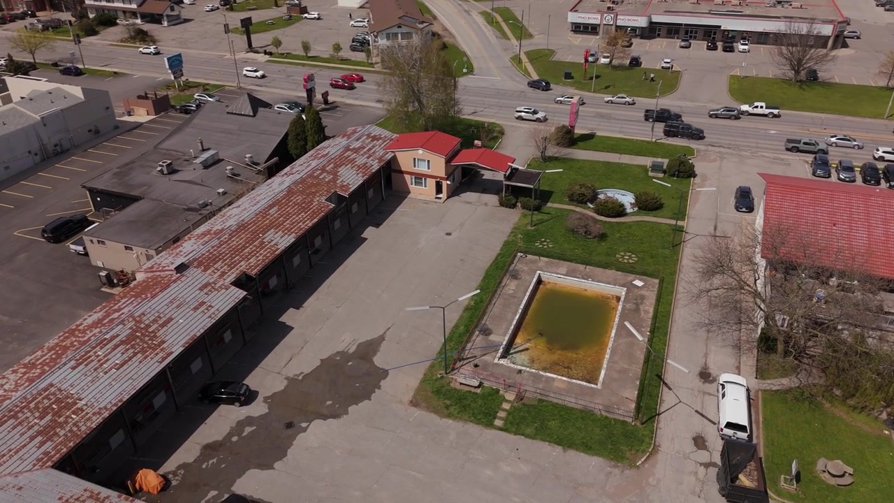 Vista drone di un motel abbandonato con piscina sporca e parcheggio vuoto, con traffico sullo sfondo.