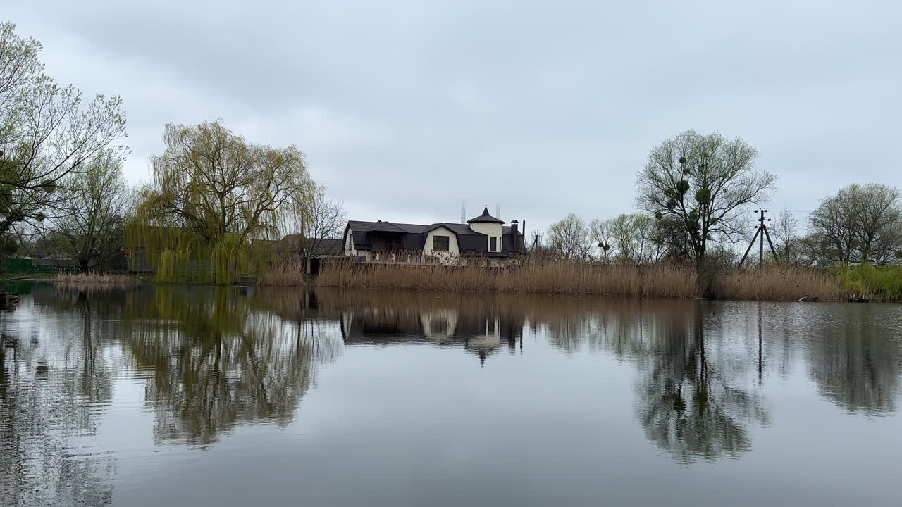 Chilly day on the calm river. Beautiful house on the bank overgrown with dry grass and bare trees in spring.