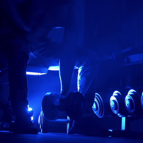 athletic man working out in gym doing exercises with dumbbells, strong male naked torso abs. blue dark light filter. selective focus.