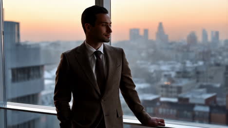 businessman in suit looks out window at city skyline during sunset