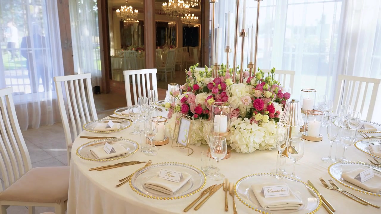 Stunning wide-angle shot of a lavishly decorated wedding reception table with lush pink and white floral centerpiece, elegant golden cutlery, crystal glassware, and soft, natural lighting.