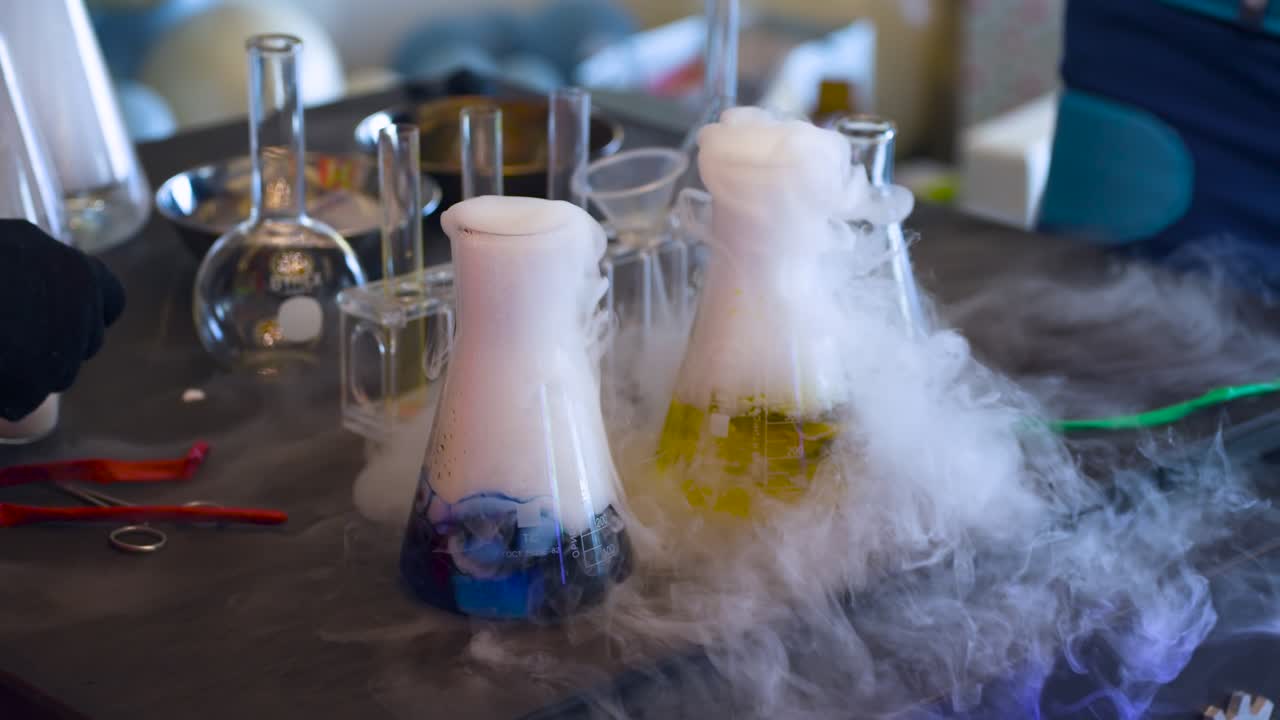 Scientifique ganté en équipement de protection orange menant une expérience à l'azote liquide avec des fioles Erlenmeyer fumantes contenant une solution chimique jaune en laboratoire.