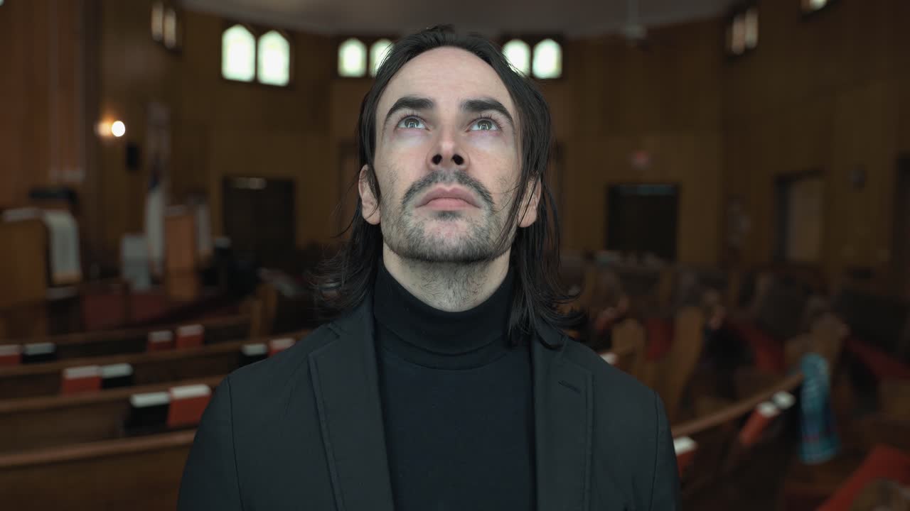 A Christian man, priest, preacher, pastor smiles in a black turtleneck and blazer stands in a church sanctuary, framed by wooden pews and soft light, creating a cinematic and contemplative mood