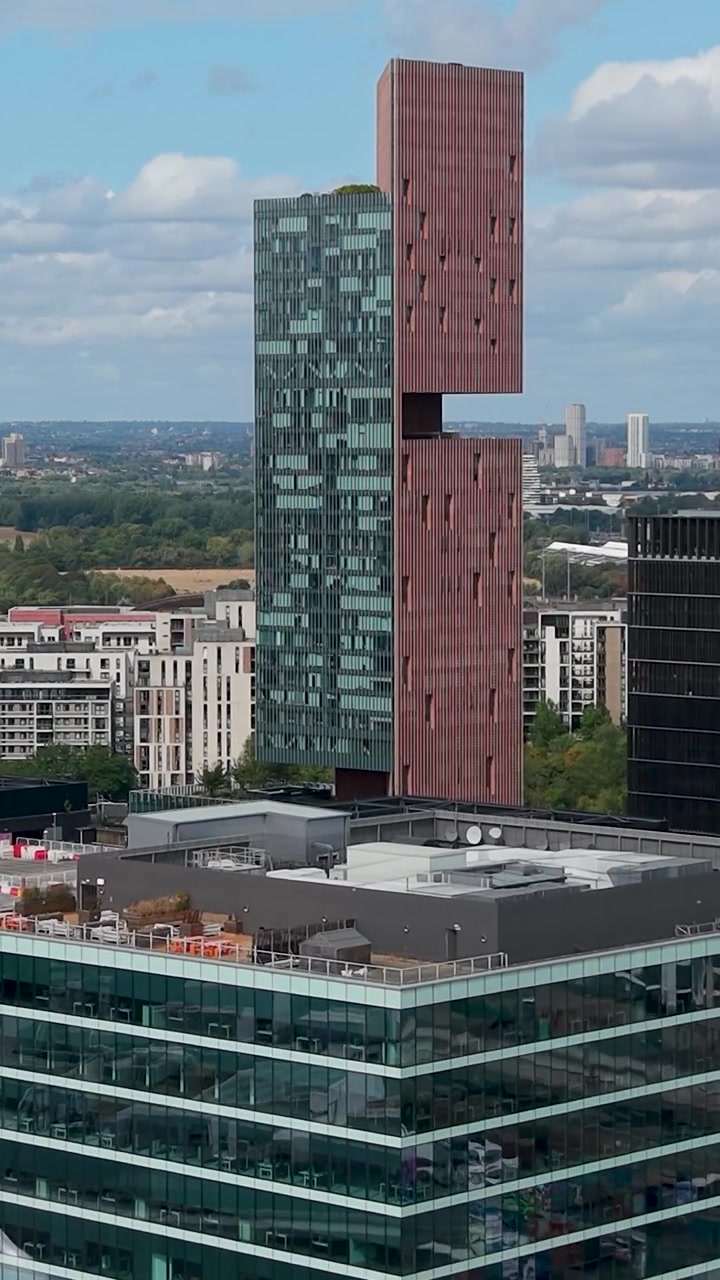 Il sorvolo aereo di Stratford Westfield si avvicina ai Manhattan Loft Gardens, rivelando lo skyline moderno di grattacieli, i tetti dei negozi e i quartieri dell'East London sotto nuvole sparse. Formato verticale