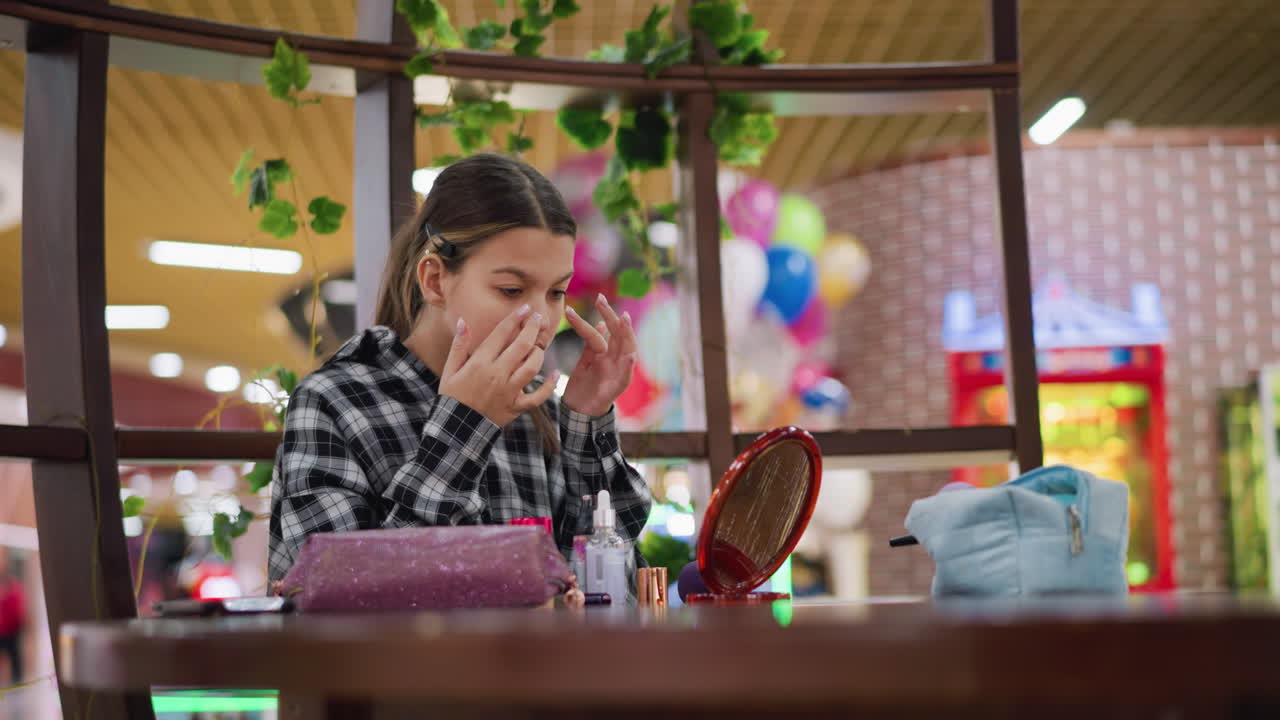 나무 테이블에 앉아 메이크업하는 카레드 셔츠를 입은 십대 소녀, 다양한 미용 제품과 액세서리가 그녀의 주위에 배열되어 있으며, 배경에는 다채로운 풍선이 있습니다.