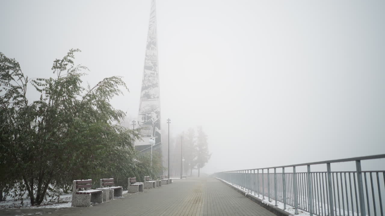 안개가 가득한 공원 벤치, 얼어붙은 나무, 철로 된 레일링, 평화로운 산책로를 따라 빛의 기둥을 은 눈이 내리는 겨울 도시 풍경, 먼 막대기가 안개의 배경에서 흐릿하게 보입니다.
