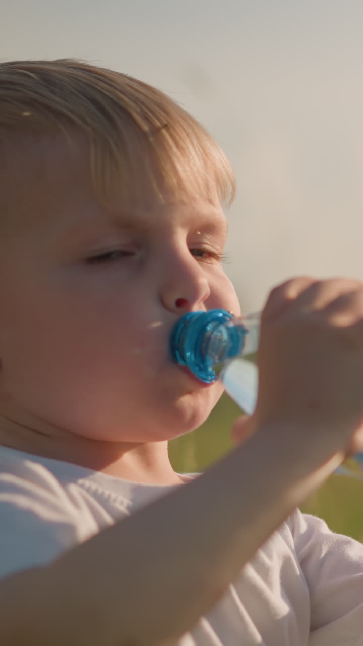 A little kid wearing a white top is sitting in a grassy field, putting a water bottle to his mouth to drink. The child appears focused and calm, enjoying a refreshing moment outdoors in the sunlight