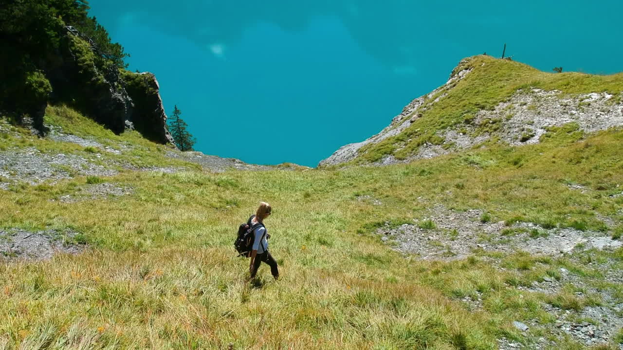 A blonde girl hikes down a Mountain at lake oeschinen in the swiss apls with the blue mountain lake in the background