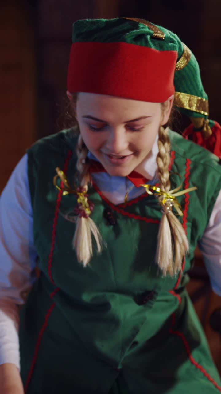 Portrait of a happy elf in the Christmas decorated room. Funny female elf in green costume with red and green hat choosing envelope among others. Vertical video
