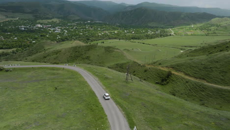 Luftaufnahme-Eines-Weißen-Fahrzeugs,-Das-Auf-Der-Straße-Mit-Blick-Auf-Die-Grünen-Hügel,-Wiesen-Und-Berge-In-Der-Nähe-Von-Aspindza-In-Georgia-Fährt