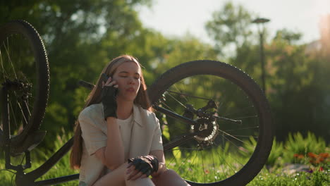 young lady sitting near her upside-down bike, engaged in phone call with a concerned and bothered expression, surrounded by lush greenery and vibrant tree