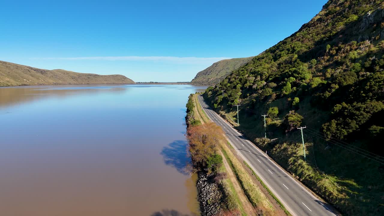 Drone footage captures a serene road alongside a tranquil lake in Akaroa, New Zealand, under clear blue skies