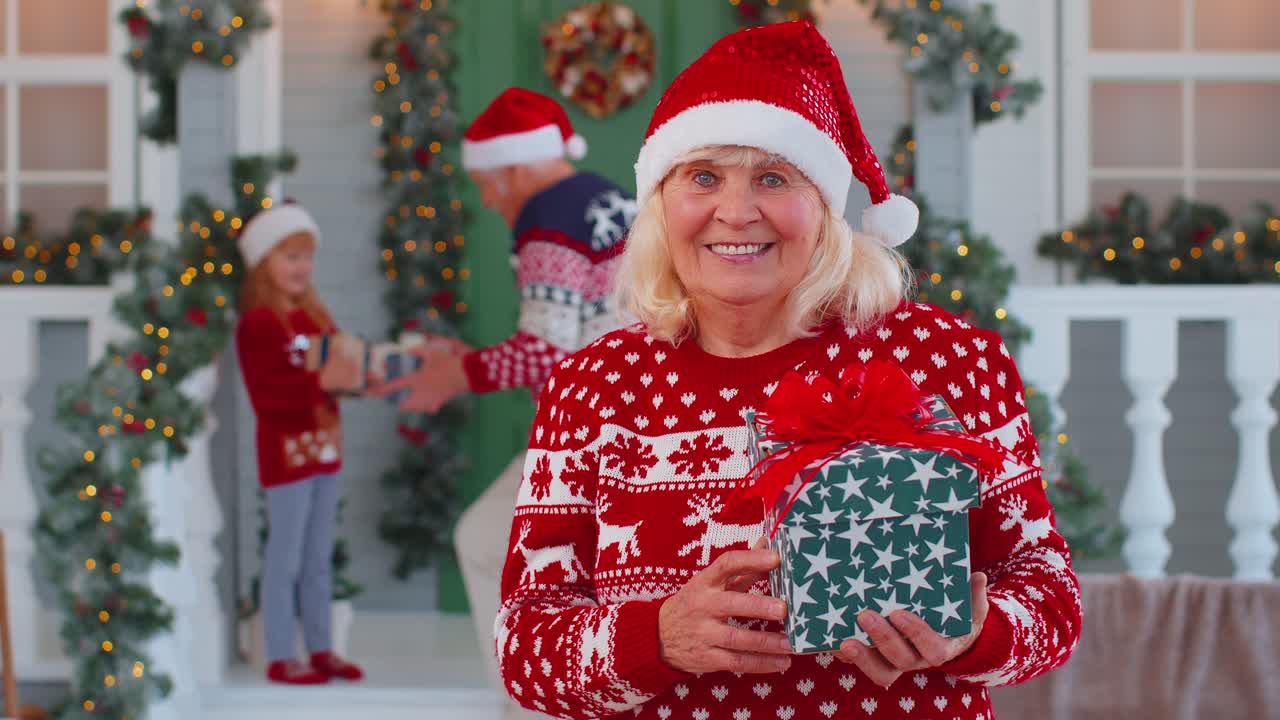 retrato de una abuela presentando una caja de regalos sonriendo cerca de una casa de navidad decorada con su familia