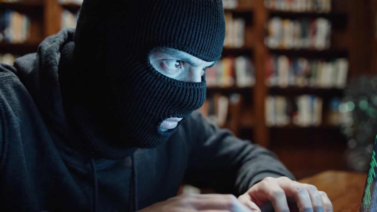 En maskeret figur fokuserer intenst på en bærbar computer, omgivet af bogreoler, hvilket illustrerer temaerne hemmeligholdelse og digitalt spionage i et akademisk miljø