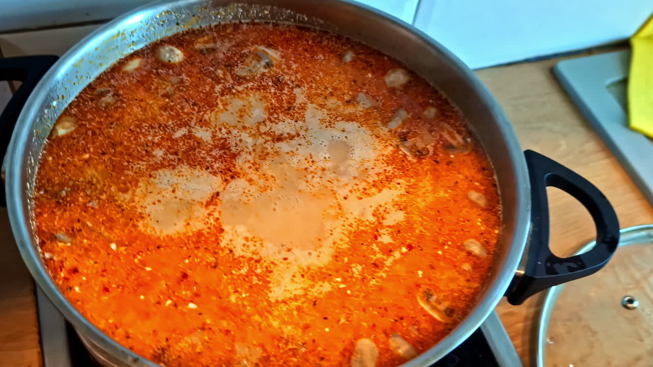 A top-down close-up shot of a large pot of traditional hot, spicy, and sour Thai Tom Yum Goong soup simmering, showing the rich red chili broth with herbs and spices