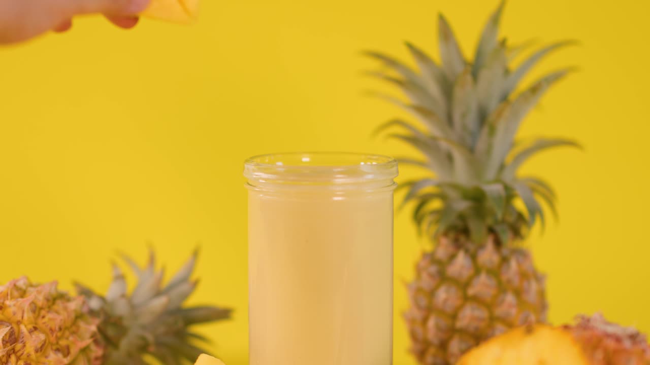 Hand places pineapple slice on smoothie glass, bright yellow background, tropical fruit, even lighting