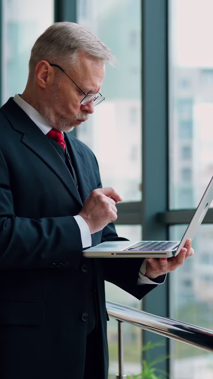 Mature senior serious businessman with laptop in hands thinking while checking e-mails standing near window with panoramic city view. Video from the side. Vertical video