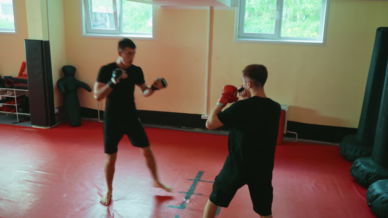 Two boxers sparring in martial arts gym wearing gloves facing each other on red mat floor fighting during training session with natural light from windows and equipment visible in background