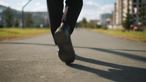 leg view of individual in black trousers and shoes walking confidently outdoors on tarred road with shadow cast on ground, surrounded by urban trees, modern buildings, and sunny blue sky