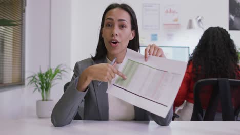 feliz mujer de negocios biracial sentada en el escritorio, sosteniendo documentos, haciendo videollamadas en una oficina moderna