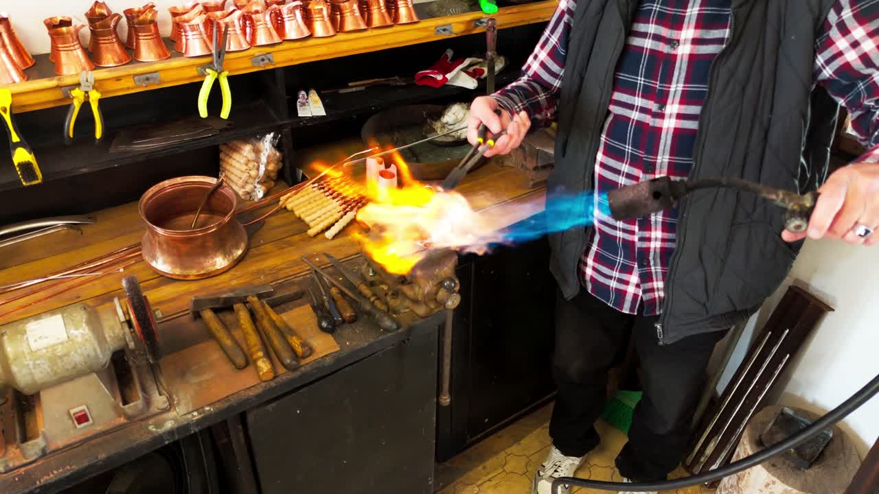 Coppersmith using blowtorch to heat copper pot in traditional workshop, part of crafting process.