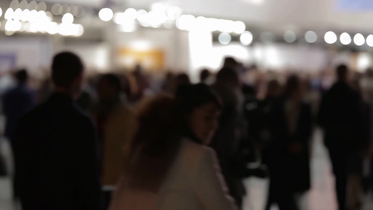 Scena sfocata di una sala eventi affollata con molti partecipanti che camminano e chiacchierano, illuminazione calda dall'alto e un'atmosfera vivace con effetto bokeh che suggerisce networking professionale e collaborazione.