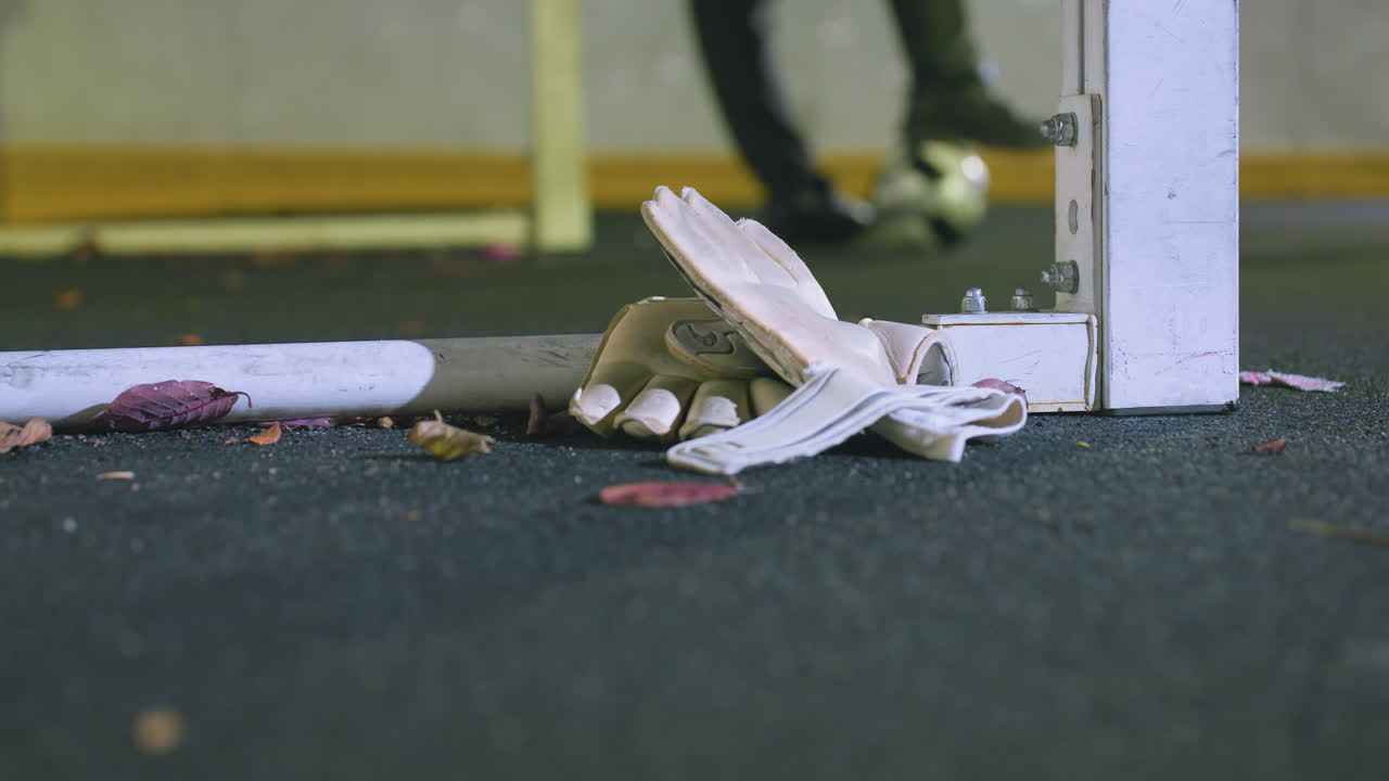 골키퍼의 장갑이 골포스트 근처에 잔디에 어져있는 잎으로 둘러싸여있는 클로즈업, 밤에 축구 공을 발 에 들고 있는 선수의 흐릿한 배경