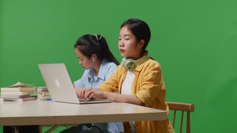 asian woman student typing on a laptop and stretching while sitting with her friend on the table in the green screen background classroom