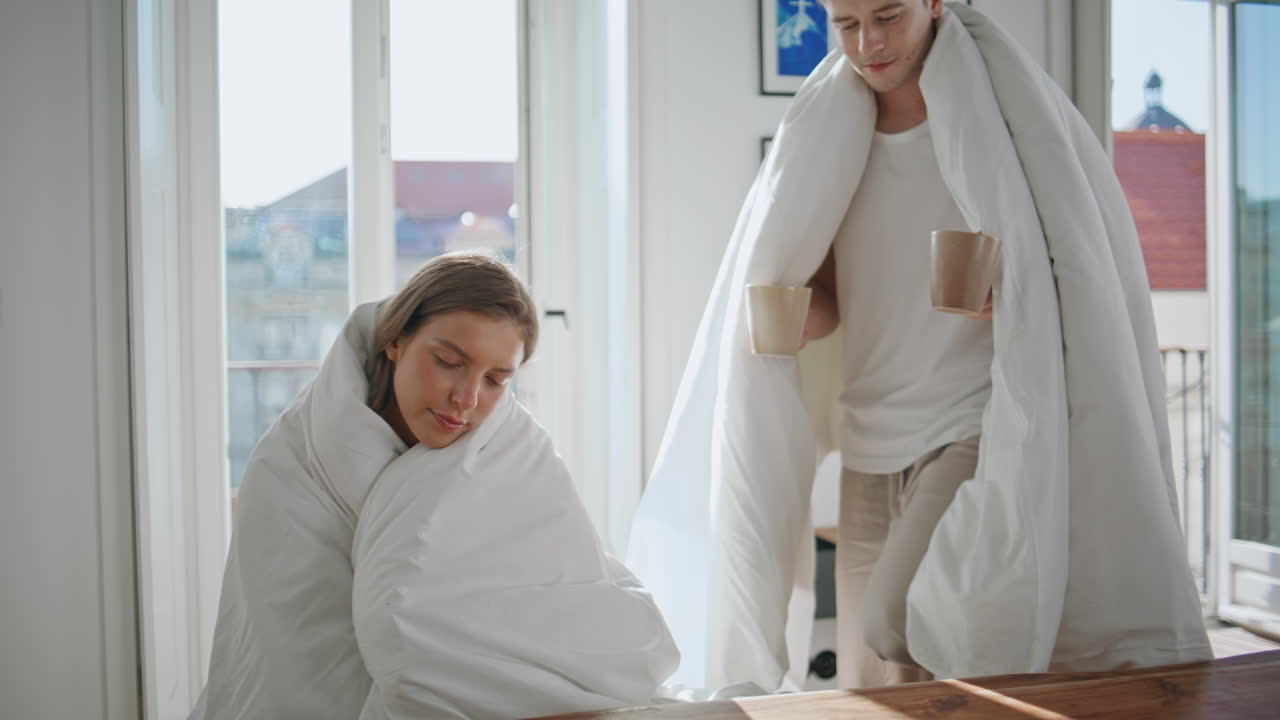 Husband handing morning coffee to sleepy wife. Cute couple with white blankets