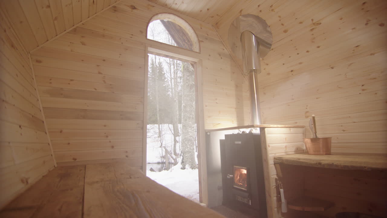 Steamy wooden sauna with fire in stove as open door shows snowy woods outside