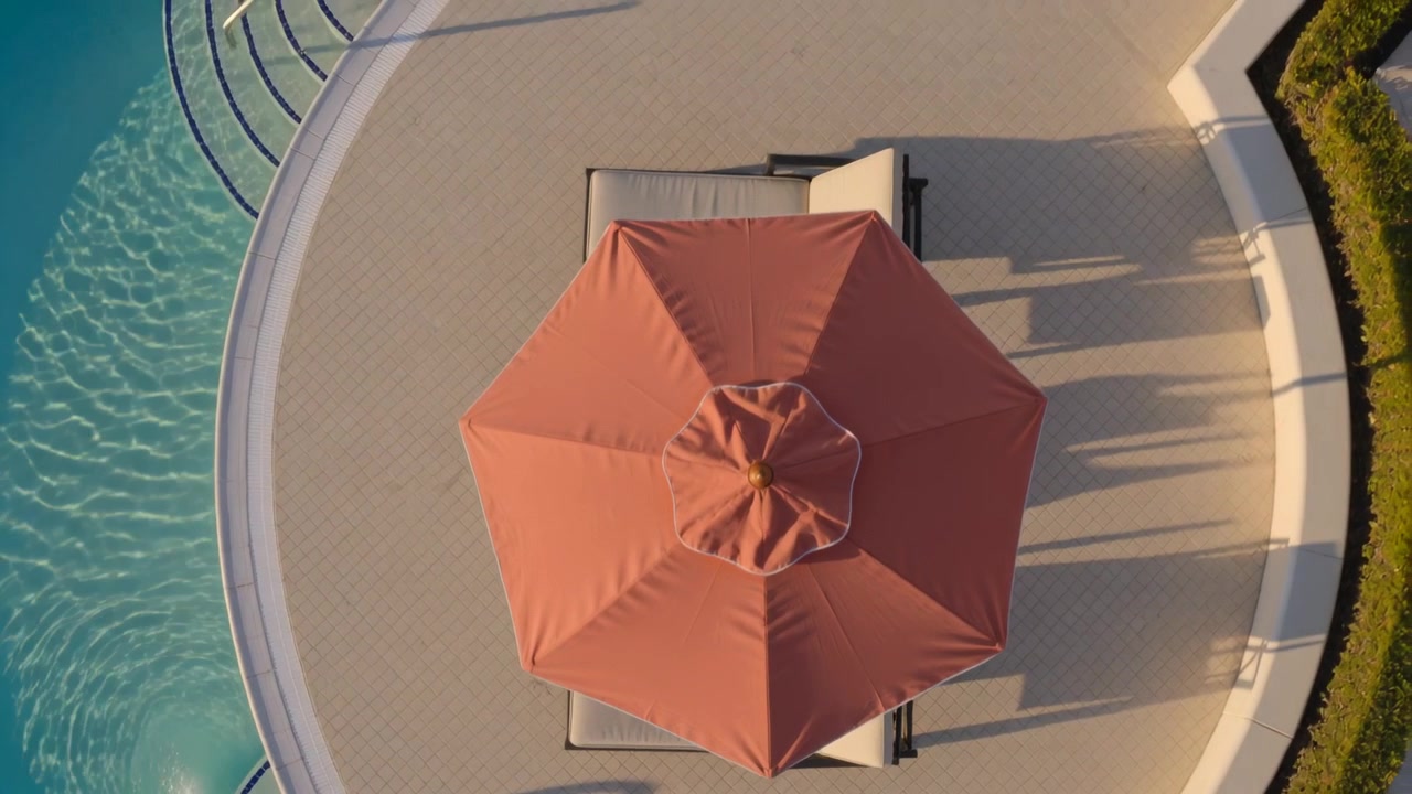 La telecamera panoramica rivela un ombrellone arancione che copre una sedia a sdraio a bordo piscina, rivelando una scaletta e una siepe.