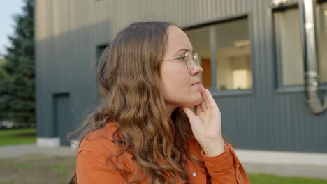 Woman-in-round-rim-glasses-scratches-chin-curious-thinking-pondering-at-sunset-in-urban-setting