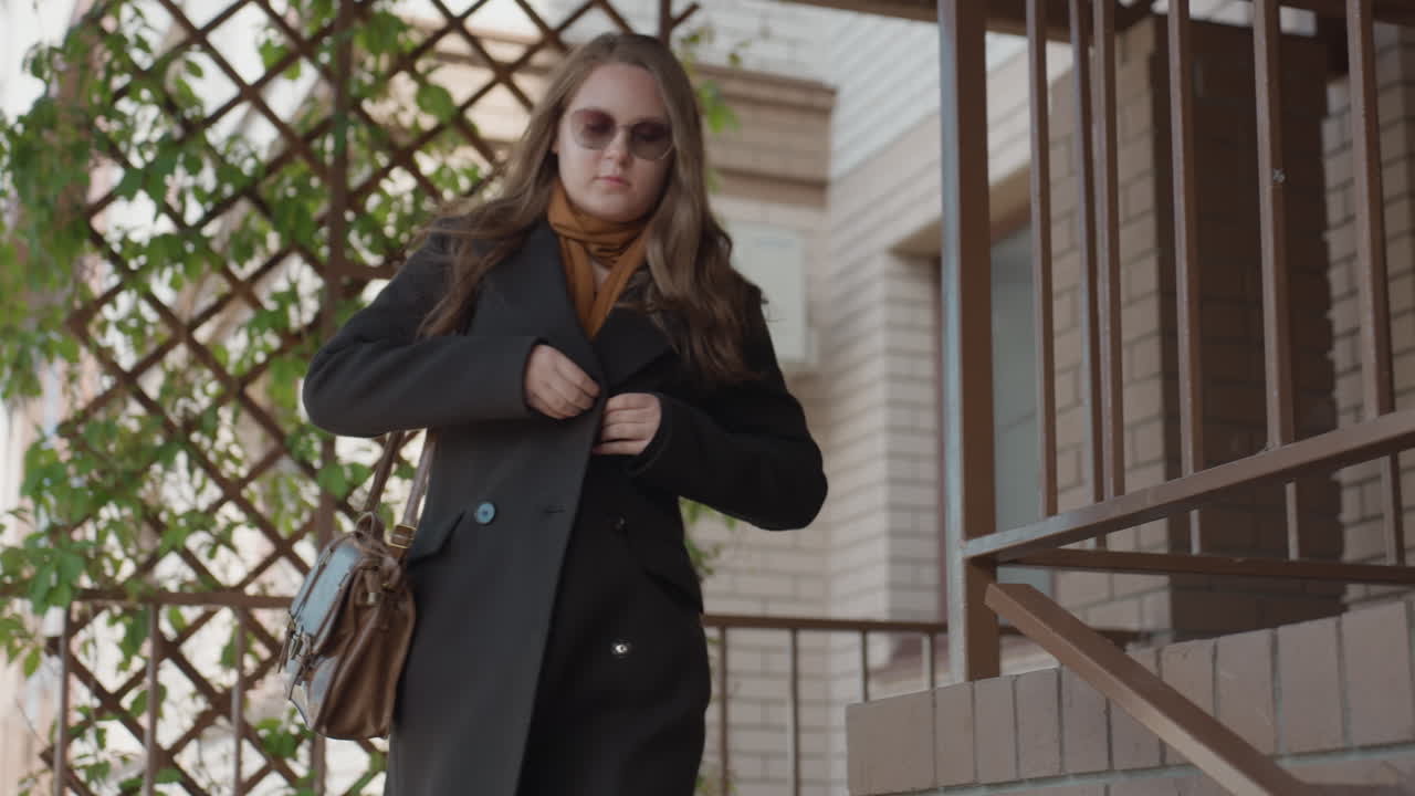 plano medio a través de barandillas de metal que muestra a un estudiante ajustándose el abrigo mientras baja unos escalones exteriores junto a una pared de bloques y flores con una escena urbana tranquila de un porche, luz del día, sutil desenfoque de movimiento, ambiente confiado