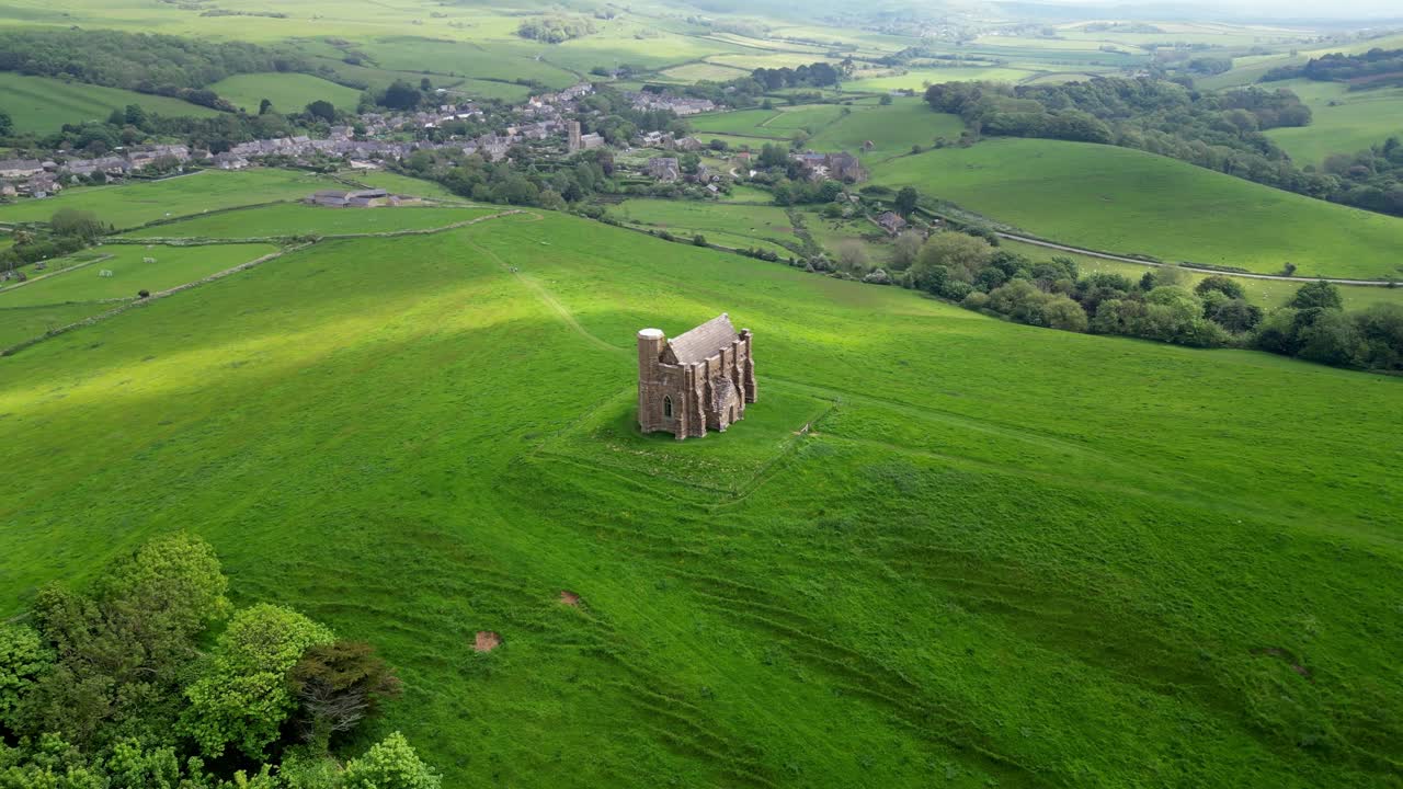 Aerial drone orbit shot of St Catherine's Chapel, Abbotsbury, Dorset, on England's South Coast with views of Chesil Beach and surrounding lush green fields. A shard of sunlight coincidentally passes.