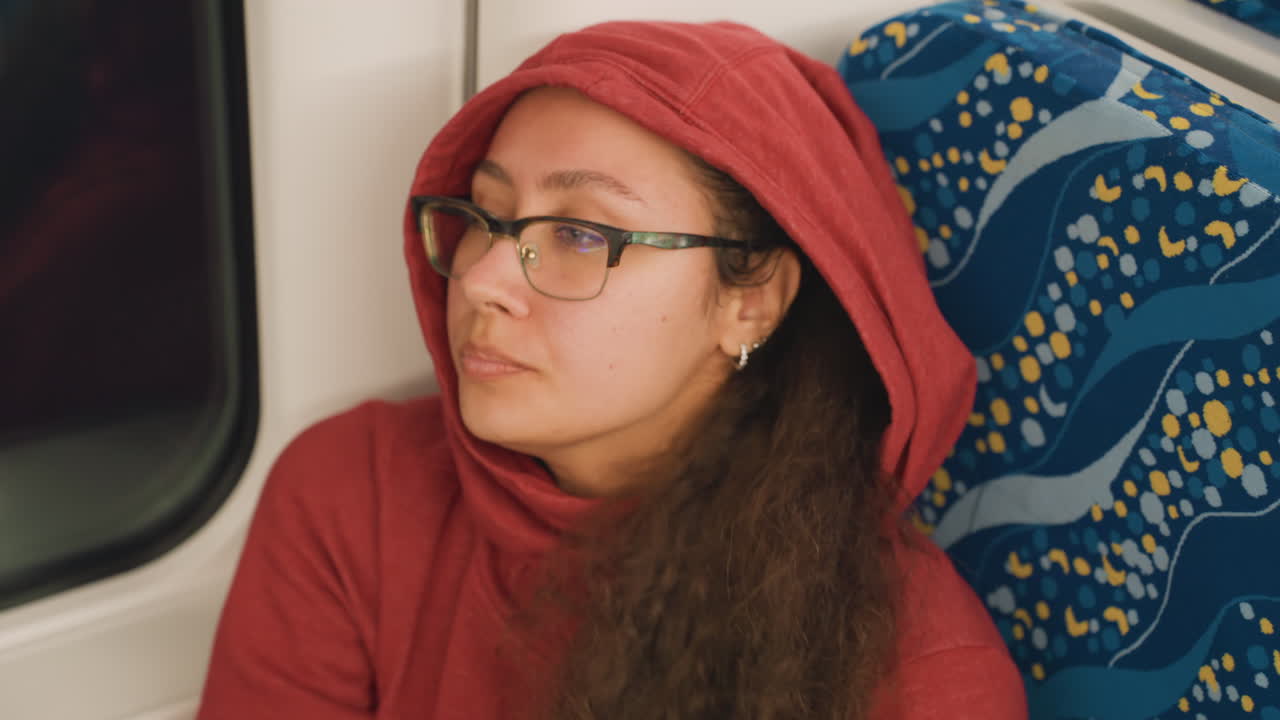 Donna che riposa con il cappuccio alzato vicino al finestrino, gli occhi si chiudono e la testa è appoggiata al sedile. Un tranquillo viaggio notturno con luce soffusa e atmosfera riflessiva. Indossa un maglione comodo, cullata dal ritmo costante delle rotaie.