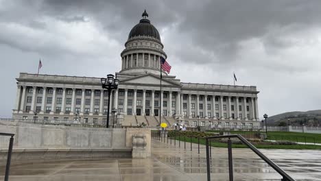 Wenn-Man-An-Einem-Bewölkten-Tag-Zur-Hauptstadt-Des-Bundesstaates-Utah-Hinaufschaut,-Sieht-Man-Menschen-Mit-Regenschirmen-Stehen-Und-Herumlaufen-Und-Die-Amerikanische-Flagge-Weht-Im-Wind