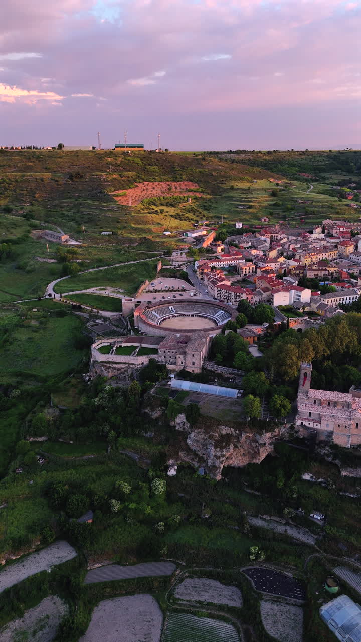 Vertical drone shot with lateral movement over Brihuega, La Alcarria, Spain. The clip shows bullring, terraced gardens, cliffs, rooftops and soft morning light over rural hills and fields