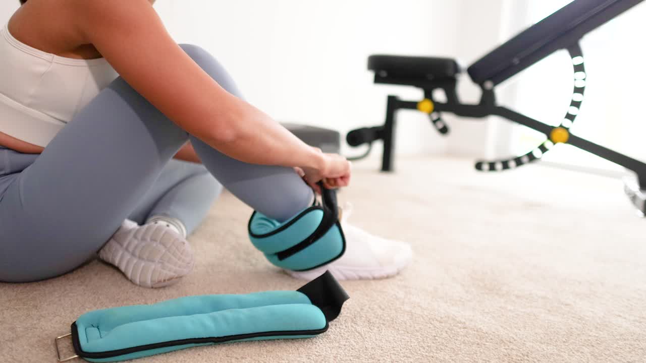 A woman in a home gym setting adjusts ankle weights, preparing for a workout. Bright lighting and neutral tones create a focused atmosphere