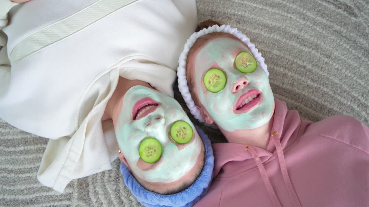 Two friends are lying on at-home carpet with mint masks and cucumber slices, breathing for spa